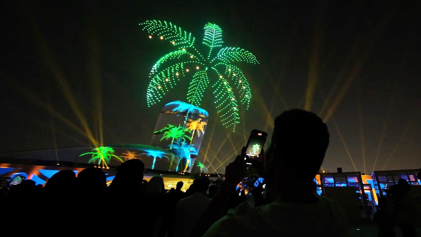 Drone night show at the Ithra Cultural Center. Hundreds of drones form a bright green palm tree in the sky, and colored images of palm trees are projected onto the building. The audience is filming the show on their phones.