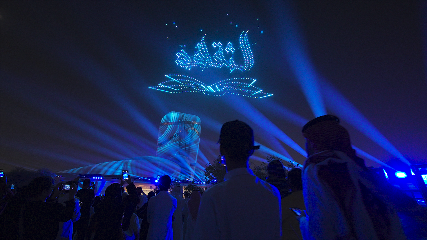 The audience watches a night light show of drones over the Ithra Cultural Center. In the sky, drones line up in the form of an open book and an Arabic inscription illuminated by blue light.