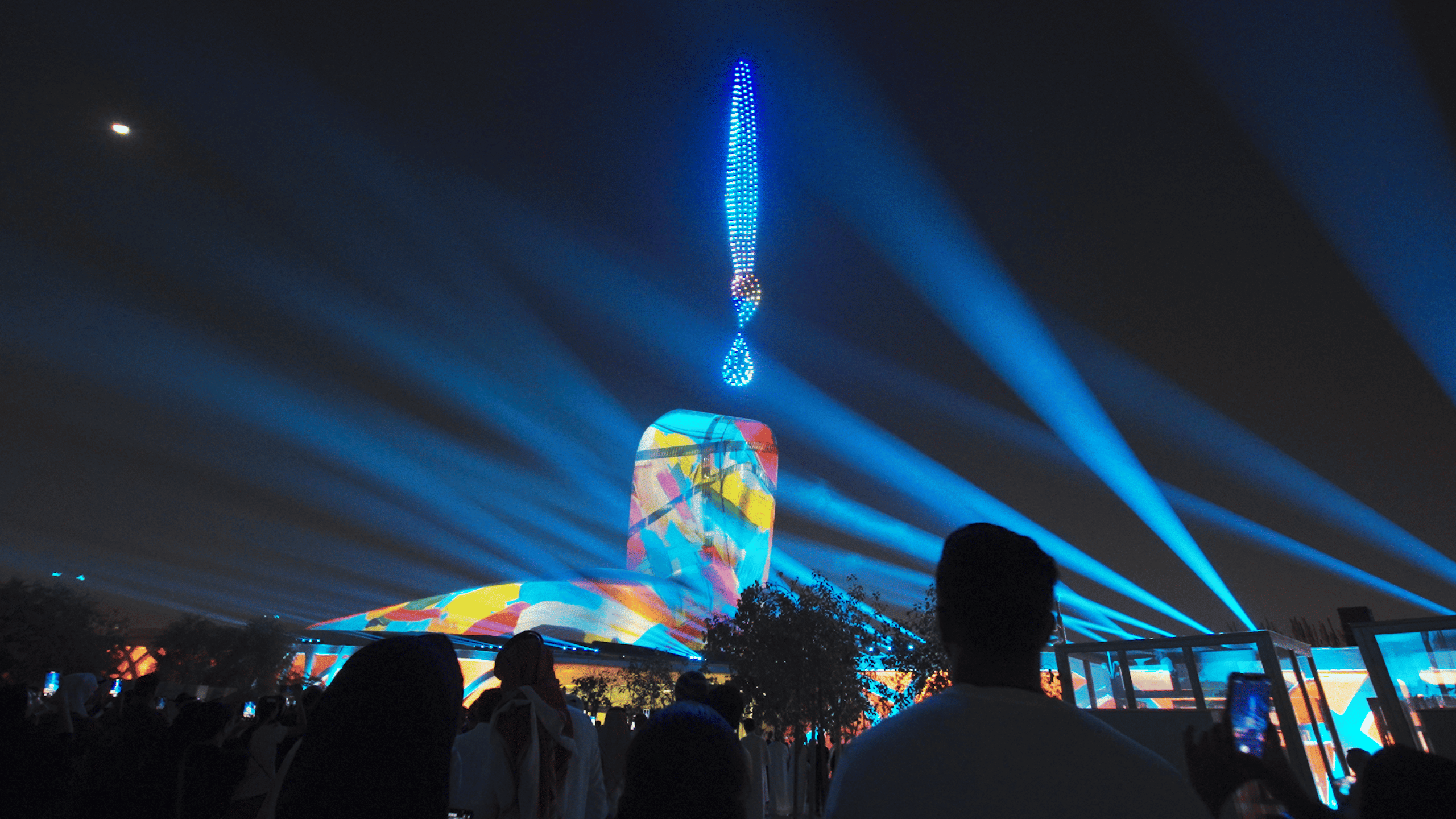 Drone night show at the Ithra Cultural Center. In the sky, hundreds of drones create an artist's brush shape with a drop of paint at the end, illuminated by blue spotlights. Bright multicolored patterns are projected on the building.