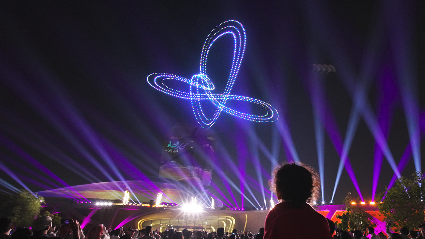 A crowd of spectators watches a night drone show at the Ithra Cultural Center. Hundreds of drones form intersecting rings of light in the sky, illuminated by purple spotlights.