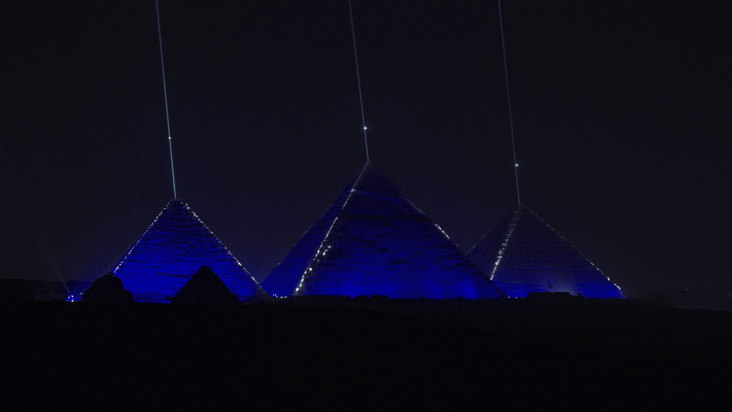 During the Lumasky drone show at the opening of the museum in Egypt, vertical rays of light rise above the pyramids, and the pyramids themselves are illuminated in deep blue at night.