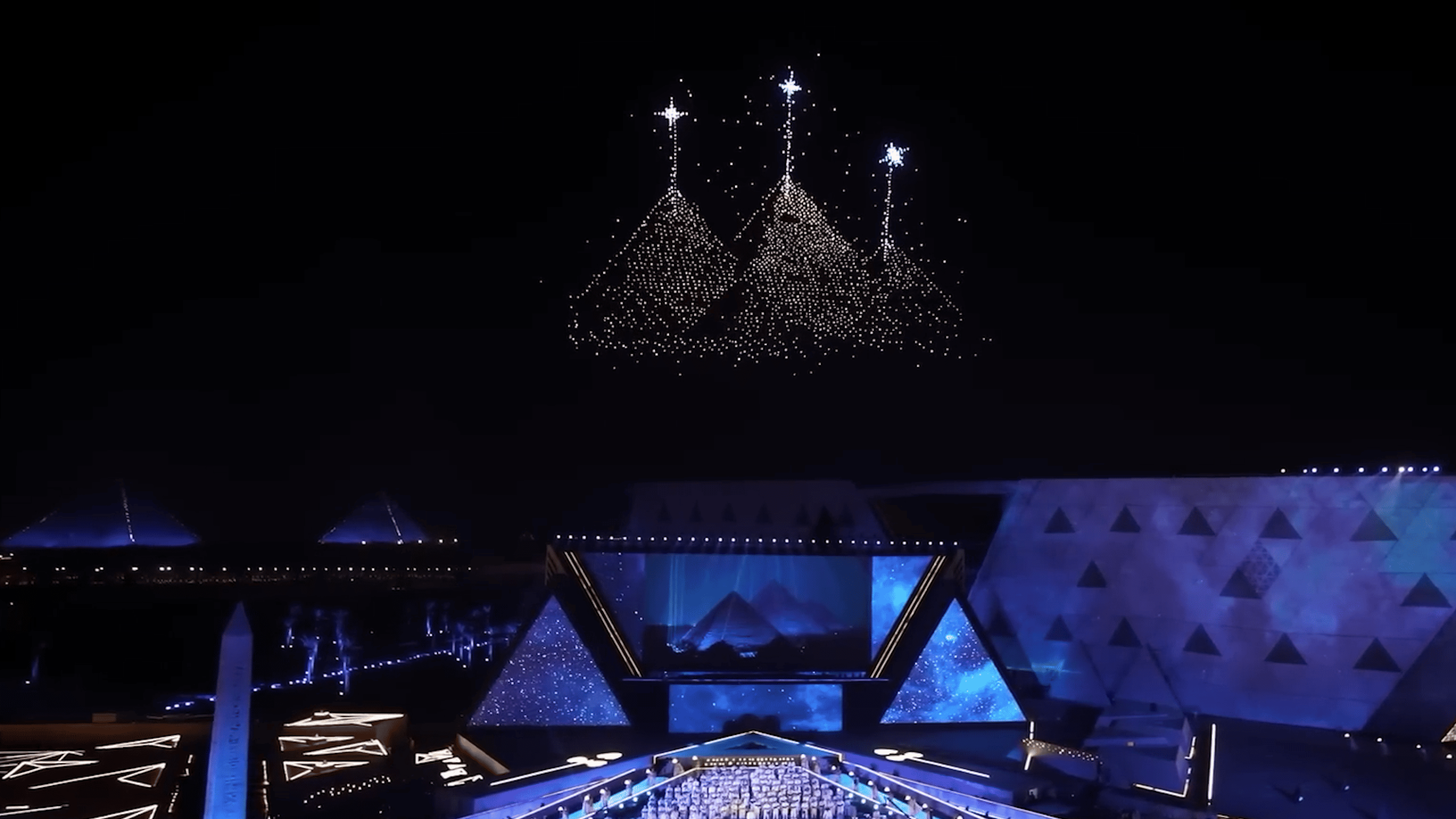 During the Lumasky drone show at the opening of the museum in Egypt, drones form a composition of three pyramids with luminous peaks in the night sky, hovering over the illuminated stage.