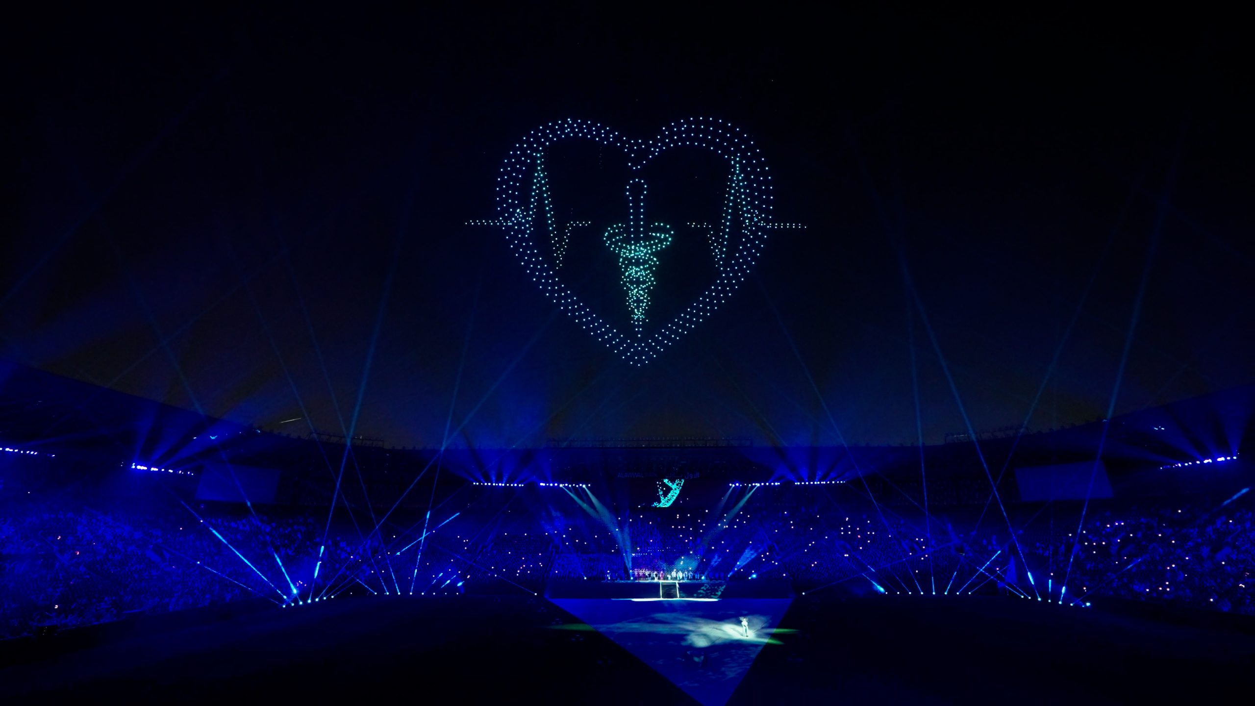 During the Lumasky Drone Show, drones form a heart with a medical symbol and a pulse line in the night sky at the “Riyadh's Doctors Got a 2,900 Drone Show for Graduation” event, above the stadium with laser effects.