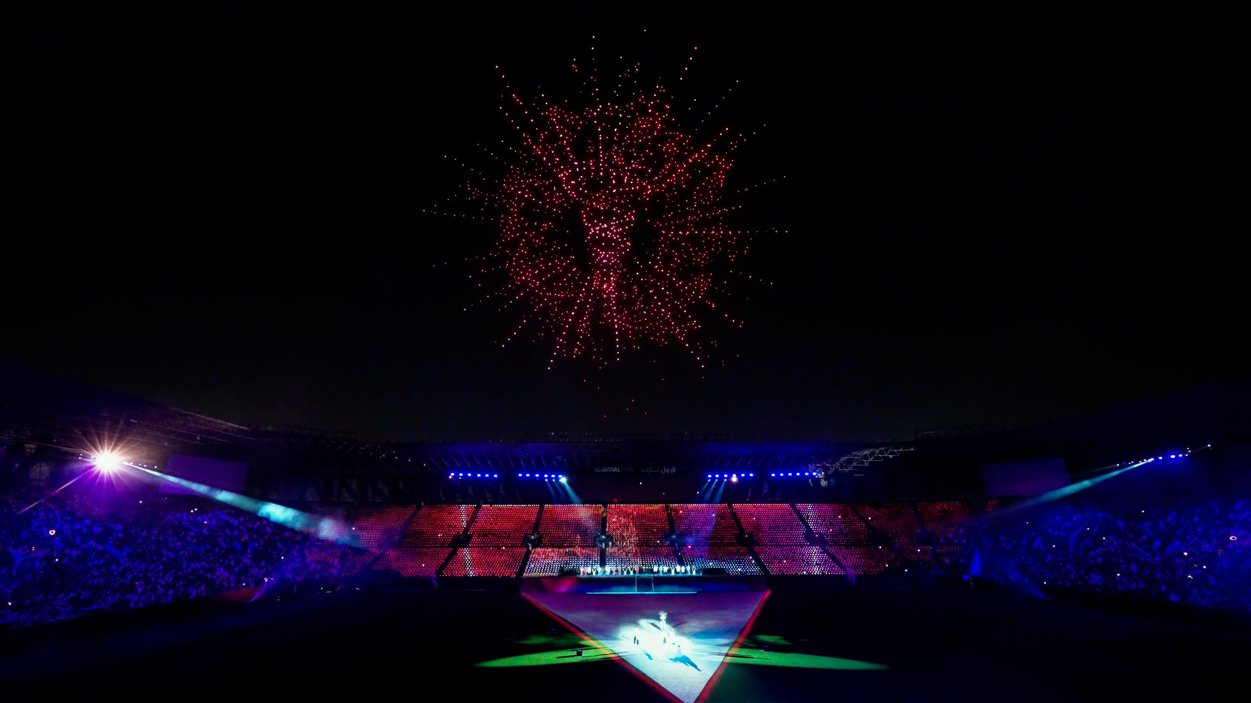 During the Lumasky Drone Show, drones form an abstract red symbol in the night sky, resembling an energy or biological structure, at the “Riyadh's Doctors Got a 2,900 Drone Show for Graduation” event, above a lighted stadium.